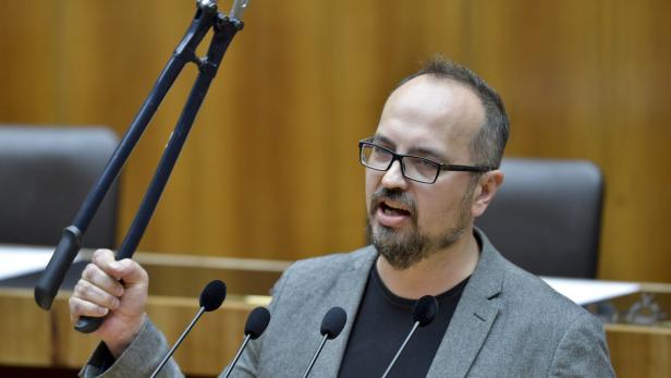 Ein Mann mit Brille hält eine Bolzenschere in die Höhe und spricht in mehrere Mikrofone.