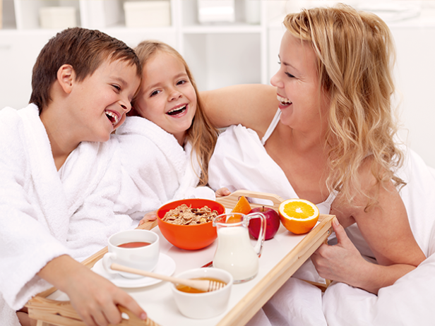 Eine Mutter und ihre Kinder lachen mit einem Frühstückstablett im Bett.