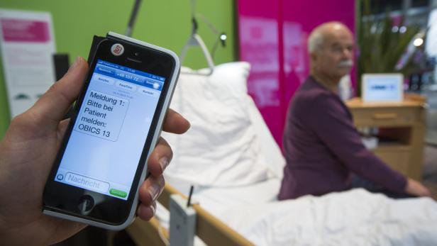 Eine Hand hält ein Smartphone mit einer Meldung, die einen Besuch bei einem Patienten anfordert. Im Hintergrund sitzt ein älterer Mann im Bett.