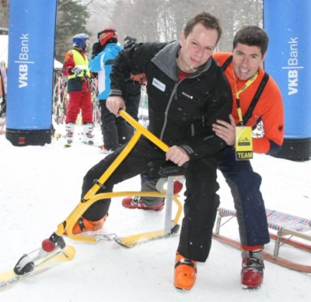 Zwei Männer posieren auf Schnee mit einem gelben Ski-Bob für ein Foto.
