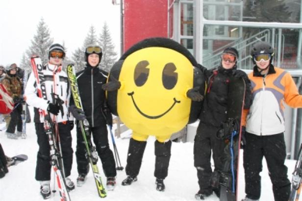 Eine Gruppe von Skifahrern posiert mit einem gelben Smiley-Maskottchen im Schnee.
