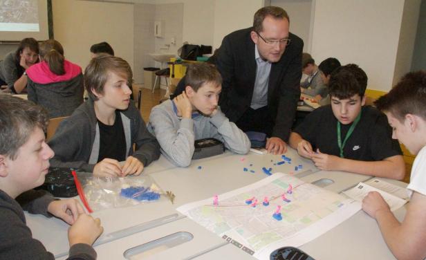Eine Gruppe von Schülern arbeitet mit einem Lehrer an einer Karte mit kleinen Figuren.