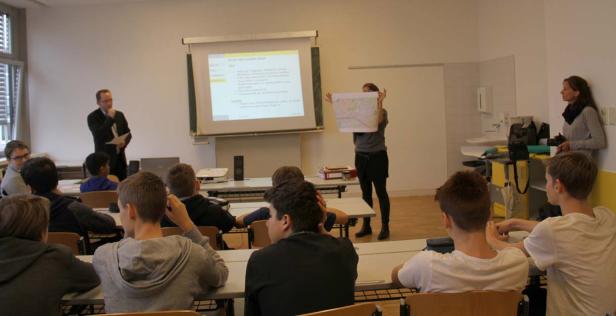 Schüler sitzen in einem Klassenzimmer, während ein Lehrer eine Präsentation hält und eine Frau eine Karte zeigt.