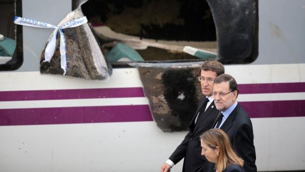Mariano Rajoy besichtigt einen beschädigten Zug, dessen Fenster mit Polizeiabsperrband gesichert ist.