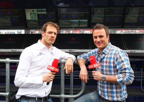 Zwei ORF-Reporter mit Mikrofonen vor Monitoren bei einer Motorsportveranstaltung.