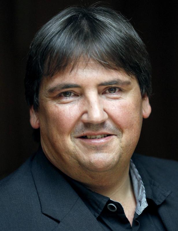 Ein Mann mit dunklen Haaren und einem schwarzen Anzug lächelt in die Kamera.