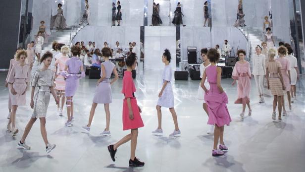 Models präsentieren pastellfarbene Kleidung und Turnschuhe auf einem Laufsteg mit einer Band im Hintergrund.