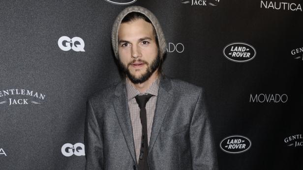 Ashton Kutcher trägt eine Mütze und ein Sakko vor einem schwarzen Hintergrund.