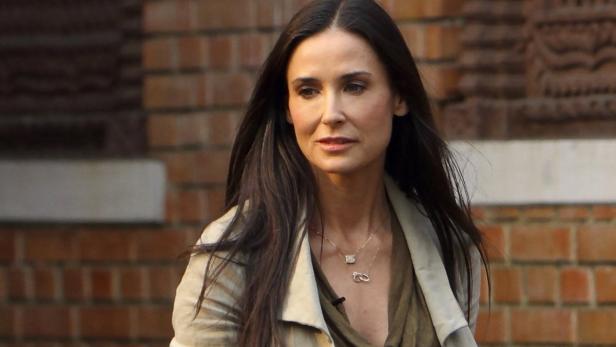 Demi Moore mit langen, dunklen Haaren und hellem Mantel vor einer Backsteinmauer.