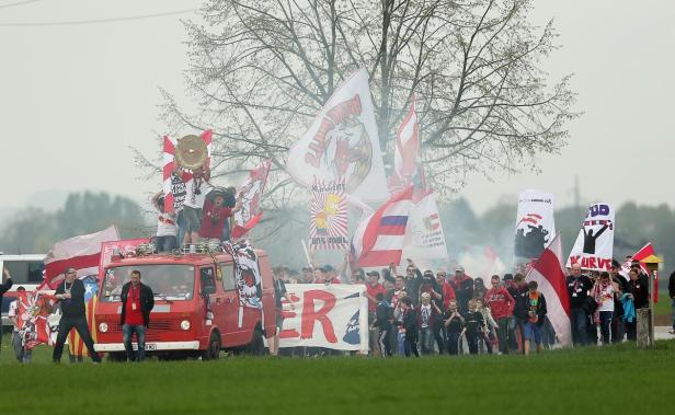 Eine Gruppe von Fußballfans zieht mit Fahnen und einem roten Lieferwagen durch eine Landschaft.