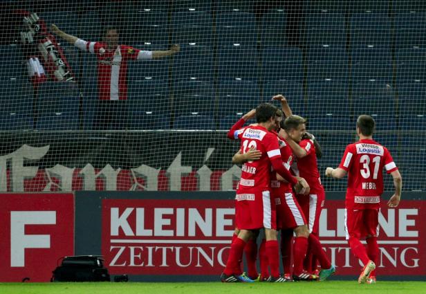 Eine Gruppe Fußballspieler in roten Trikots feiert ein Tor vor einem Fan im Hintergrund.