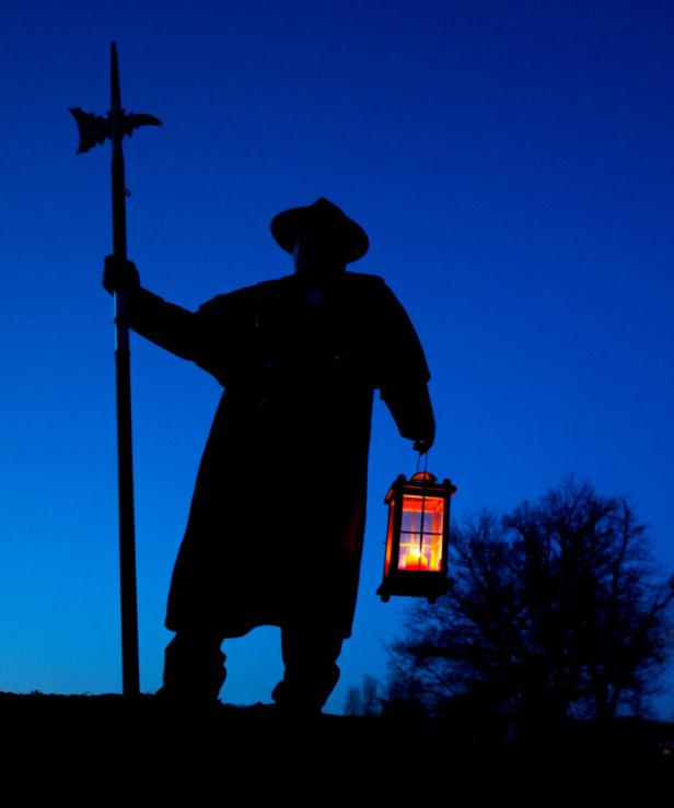 Eine dunkle Silhouette einer Person mit Laterne und Hellebarde vor blauem Himmel.