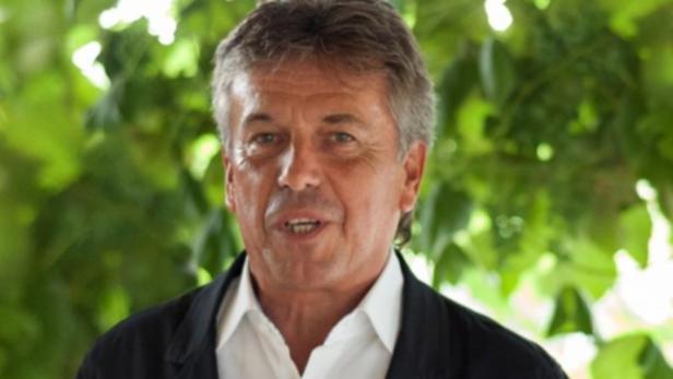 Ein Mann mit grau meliertem Haar und einem schwarzen Sakko vor grünem Laub.