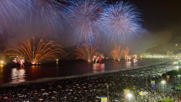 Eine Menschenmenge feiert am Strand mit einem farbenfrohen Feuerwerk über dem Meer.