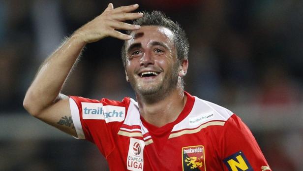 Ein lächelnder Fußballspieler in rotem Trikot wischt sich den Schweiß von der Stirn.