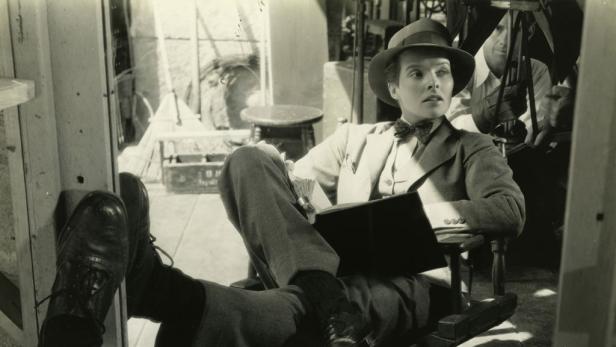 Katharine Hepburn entspannt in einem Hosenanzug und Hut am Filmset.