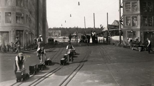 Männer bewegen Miniaturautos auf Schienen vor bemalten Stadtkulissen.