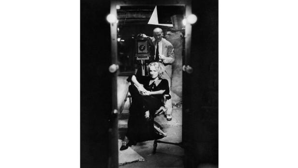 Marlene Dietrich sitzt vor einer Kamera, die von einem Fotografen bedient wird.