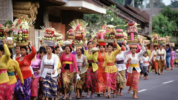 Eine Prozession von Frauen in traditioneller Kleidung trägt Opfergaben auf dem Kopf.