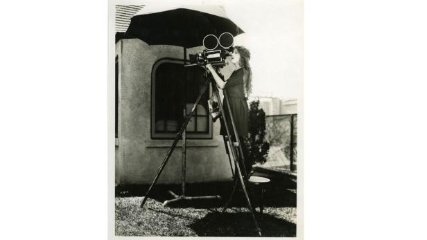 Eine Frau mit langen Haaren bedient eine Filmkamera auf einem Stativ im Freien.