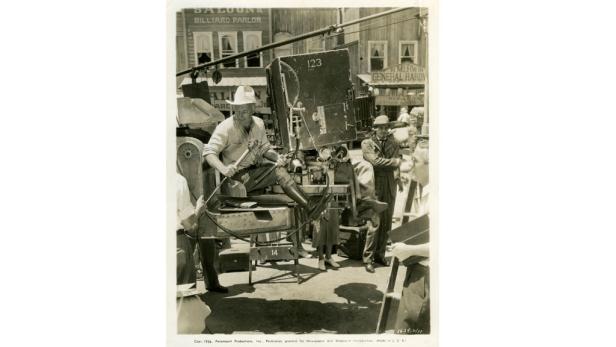 Ein Mann mit Hut sitzt auf einer Filmkamera vor einer Westernkulisse.