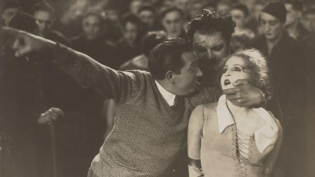 Szene mit einer bedrohten Frau, umringt von Männern, einer zeigt auf etwas im Hintergrund.