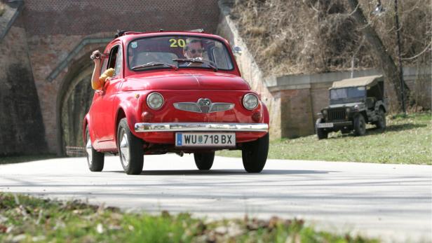 Ein roter Oldtimer fährt auf einer Straße, während eine Person aus dem Fenster filmt.