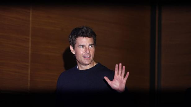 Tom Cruise winkt vor einem holzvertäfelten Hintergrund.