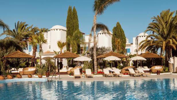 Ein Swimmingpool vor einem Hotelresort mit Liegestühlen und Sonnenschirmen.