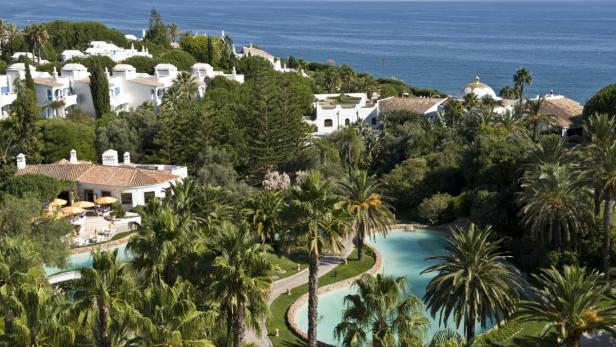 Blick auf eine luxuriöse Ferienanlage mit weißen Gebäuden, Palmen und einem Pool am Meer.