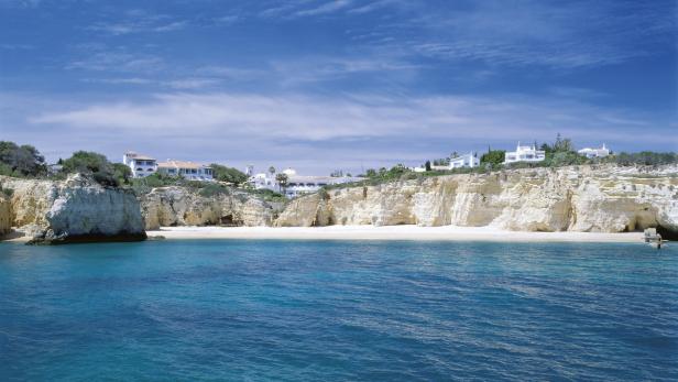 Blick auf einen Strand mit Klippen und weißen Gebäuden unter blauem Himmel.