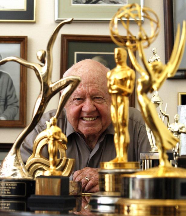 Mickey Rooney lächelt hinter einer Sammlung von Auszeichnungen, darunter ein Oscar und ein Emmy.