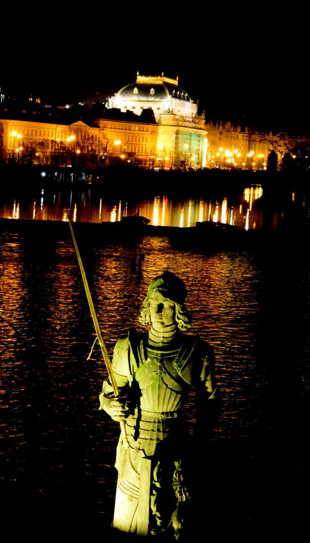 Eine Statue eines Ritters mit Schwert vor der beleuchteten Prager Altstadt bei Nacht.