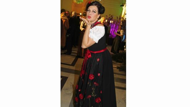 Eine Frau in Tracht mit rotem Mieder und Mohnblumen auf dem Rock sendet einen Kuss.