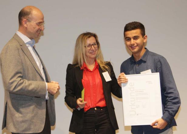 Ein junger Mann hält eine Urkunde in den Händen, flankiert von zwei Personen.