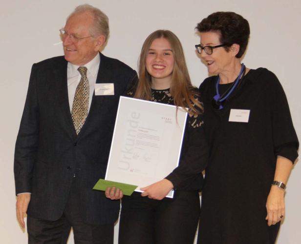 Eine junge Frau hält eine Urkunde, flankiert von zwei älteren Personen.