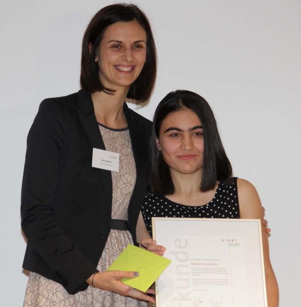 Zwei Frauen posieren mit einer Urkunde und einem Umschlag vor einer weißen Wand.