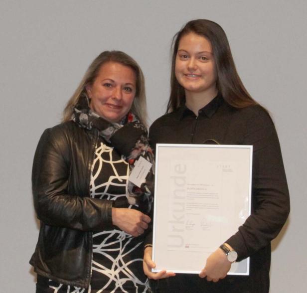 Zwei Frauen posieren mit einer Urkunde vor einer hellen Wand.