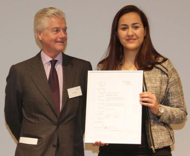 Eine junge Frau hält eine Urkunde der START-Stiftung neben einem Mann im Anzug.