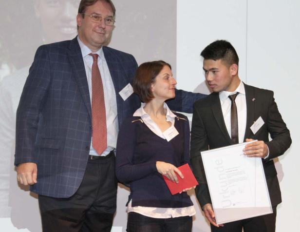 Ein Mann hält eine Urkunde, während eine Frau und ein anderer Mann daneben stehen.