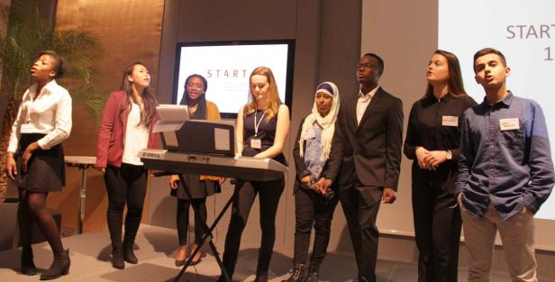 Eine diverse Gruppe junger Leute singt gemeinsam vor einem Keyboard auf einer Bühne.