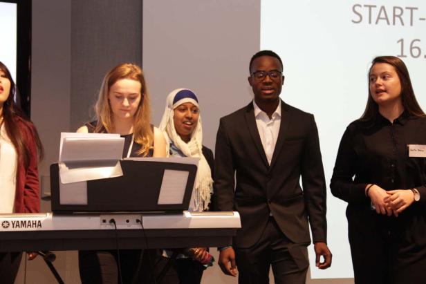 Eine Gruppe junger Leute steht an einem Keyboard und singt vor einer Leinwand.