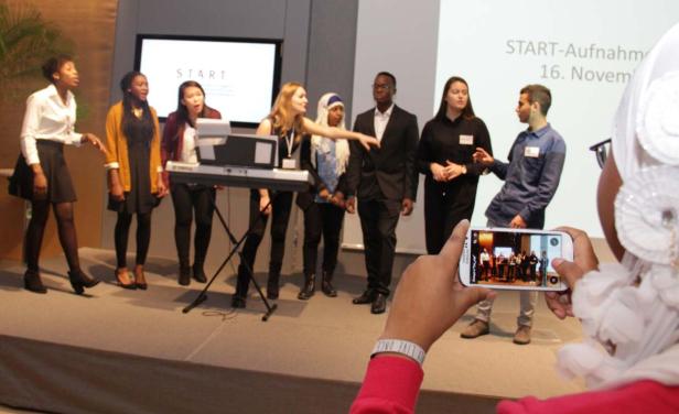 Eine Gruppe junger Leute steht auf einer Bühne und singt, während eine Person ein Foto mit ihrem Handy macht.