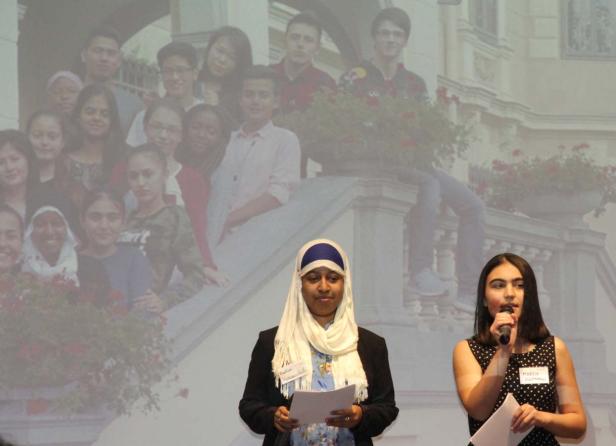 Zwei junge Frauen halten Reden vor einer Projektion einer Gruppe von Jugendlichen.