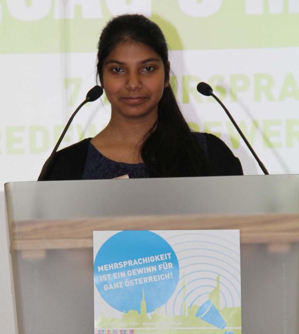 Eine junge Frau steht an einem Rednerpult mit dem Slogan „Mehrsprachigkeit ist ein Gewinn für ganz Österreich!“.