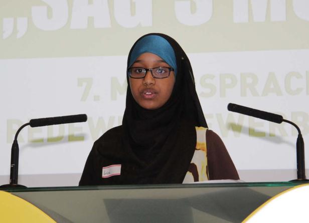 Eine junge Frau mit Brille und Hijab spricht bei einer Veranstaltung am Rednerpult.