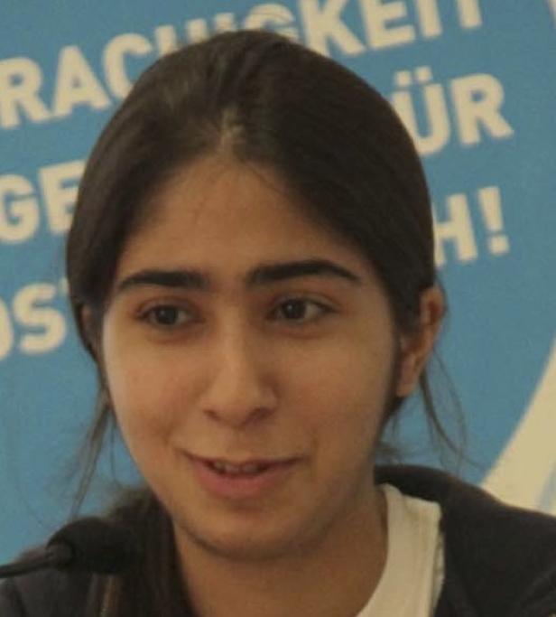 Eine junge Frau mit dunklen Haaren lächelt vor einem blauen Hintergrund.