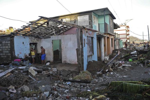 Ein zerstörtes Haus mit Trümmern und zwei Personen davor.