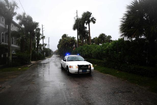 Ein Polizeiauto fährt auf einer regnerischen Straße mit Palmen und Häusern im Hintergrund.