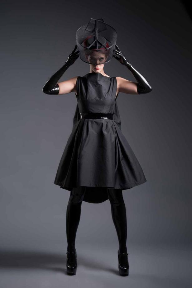 Eine Frau in einem schwarzen Kleid, Leggings und Handschuhen posiert mit einem extravaganten Hut.
